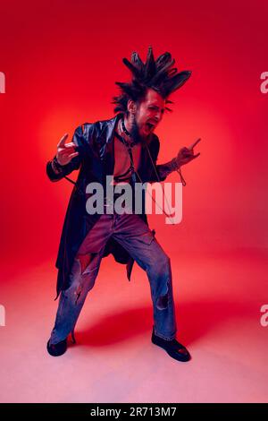 Porträt eines jungen Punk in außergewöhnlicher Kleidung, Frisur und Make-up vor rotem Studiohintergrund in Neonlicht Stockfoto