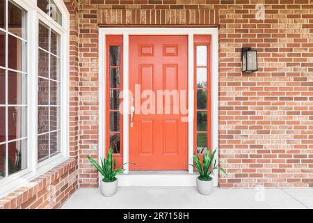 Die rote Haustür eines Hauses ist von roten Ziegeln und Pflanzen umgeben, die vor den Fenstern sitzen. Stockfoto