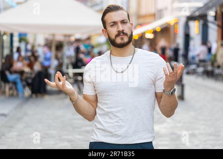 - Was? Was? Warum? Ein überraschter, frustrierter, bärtiger junger Mann, der sich die Hand hebt und nach dem Grund des Scheiterns fragt, der Ungläubigkeit zeigt, die durch Unruhen im Freien verursacht wird. Ein verwirrter Kerl, der in der Sunshine Street der Stadt spaziert Stockfoto