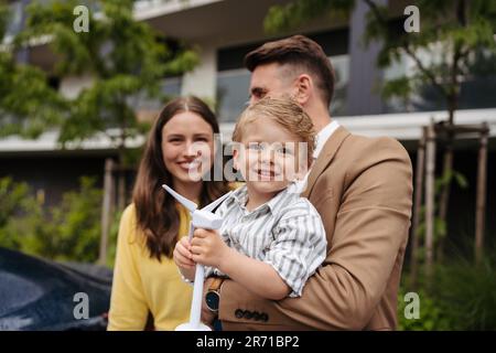 Portriat des süßen Jungen hält Windturbine, während Vater sein Elektroauto auf der Straße auflädt. Stockfoto