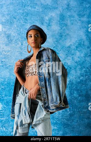 Moderne afroamerikanische Frau mit auffälligem Make-up und Baskenmütze, die eine Denim-Jacke, ein Oberteil mit Tiermuster und Jeans trägt und auf dem blau strukturierten Backgro steht Stockfoto