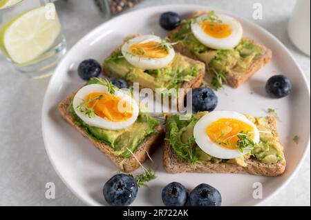 Toast zum Frühstück mit Avocado-Aufstrich, gekochten Eiern und Kresse auf einem Teller. Serviert mit einem Glas Wasser und Kaffee auf einem Küchentisch. Stockfoto