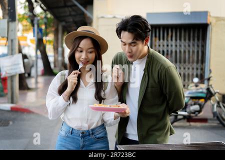 Asiatischer Paarreisender mit legerer Kleidung und Hut in einer Beziehung, der am Abend auf dem Street Food-Markt einen Dessert-Snack kauft. Rucksacktourist in Bangkok, Thailand. Stockfoto