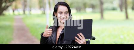 Lächelnde Frau, die im Rollstuhl im Park sitzt und den Daumen auf dem Tablet der Kamera zeigt Stockfoto