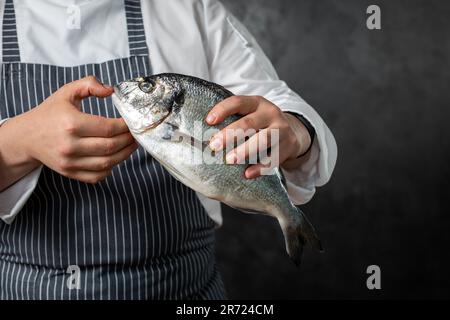 Ein anonymer männlicher Fischhändler in einer gestreiften Schürze, der frischen, ungekochten Fisch vor schwarzem Hintergrund hält Stockfoto