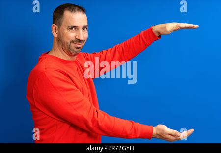 bärtiger hispanischer Mann in einem roten Pullover mit offenen Armen im Profil, der etwas bietet, während er vor der Kamera lächelt, isoliert vor blauem Hintergrund. Stockfoto