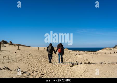 Ein paar Reisende mit einem Rucksack, Hand in Hand, spazieren entlang der sandigen Dead Dunes, an der Kurischen Nehrung, am Ufer der Ostsee. Stockfoto