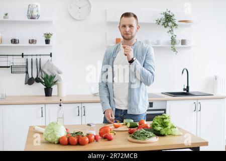 Attraktiver junger Mann in Jeansshirt, der am Esstisch posiert, mit Bio-Produkten und Blattgrün oben drauf. Ein gutaussehender Mann, der kulinarische Messerkenntnisse beherrscht, während er vegetarischen Salat drinnen zubereitet. Stockfoto