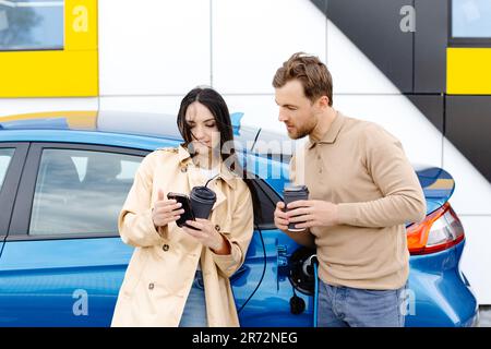 Ein junges Paar, das mit einem Elektroauto unterwegs ist und an der Ladestation hält. Freund, der das Kabel zum Aufladen einsteckt. Ein Mann, der mit einer Freundin redet Stockfoto