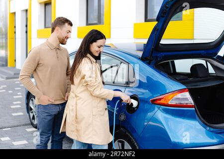 Ein junges Paar, das mit einem Elektroauto unterwegs ist und an der Ladestation hält. Eine Freundin, die das Ladegerät hält, das Auto ansieht und lächelt. Stockfoto