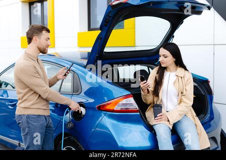 Ein junges Paar, das mit einem Elektroauto unterwegs ist und an der Ladestation hält. Freund, der das Kabel zum Aufladen einsteckt. Ein Mann, der mit einer Freundin redet Stockfoto