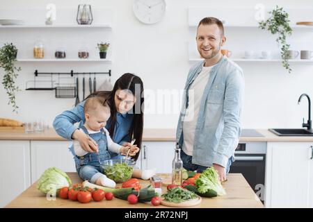 Wunderschöne Mutter und Tochter, die mit dem Mischen von Zutaten in der Schüssel beschäftigt sind, während der glückliche Vater beiseite lächelt. Fröhlicher, gutaussehender Vater, der die Vorteile der Zusammenarbeit mit der Familie in der Küche genießt. Stockfoto