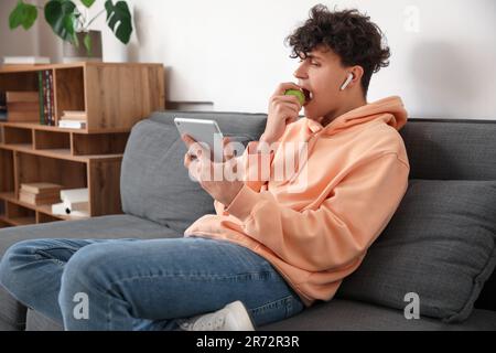 Männlicher Student mit Tablet-Computer, der zu Hause Apfel auf dem Sofa isst Stockfoto