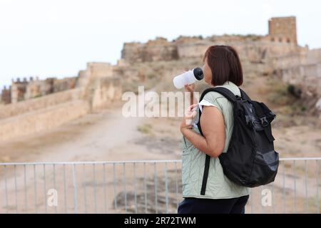 Erwachsene Reisende Frau mit Rucksack trinkt Wasser, während sie sich nach den Spaziergängen in den Ruinen der alten europäischen Festung im Freien entspannen. Rentner summieren sich Stockfoto