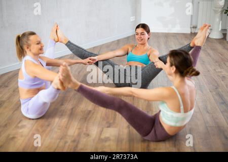 Lächelnde junge Frauen, die auf Mehl sitzen, Hände halten und sich gegenseitig die Füße berühren, nachdem sie im Fitnesscenter trainiert haben Stockfoto
