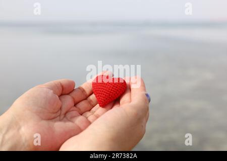 Seereise und Liebe am Sandstrand, rotes Strickherz in weiblichen Händen am Wasser. Valentinstag, Konzept einer romantischen Reise Stockfoto