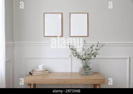 Zwei leere vertikale Bildmodelle, die an der Wand hängen. Eine Tasse Kaffee, Bücher. Holzschreibtisch, Tisch. Vase, grünes Gras und KuhPetersilie. Minimal Stockfoto