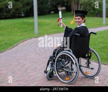 Glückliche weiße Frau im Rollstuhl dreht sich um und hält ihr Diplom draußen. Stockfoto