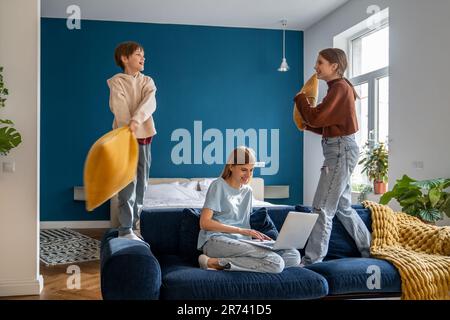 Mutter, die an einem Laptop arbeitet, während ihre aktiven, lauten Kinder mit Kissen spielen und Spaß haben Stockfoto