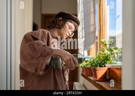 Hipstermädchen gießt Hauspflanzen, während sie zu Hause am Fenster steht Stockfoto