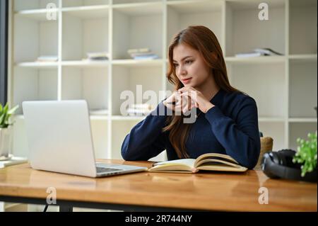 Eine inspirierte junge asiatische Geschäftsfrau oder Studentin konzentriert sich darauf, auf ihren Laptop-Bildschirm zu schauen, einen Online-Artikel zu lesen und an ihr zu arbeiten Stockfoto