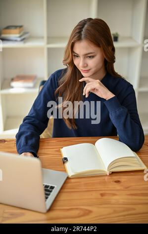 Eine charmante und inspirierte junge asiatische Geschäftsfrau oder Studentin konzentriert sich darauf, auf ihren Laptop-Bildschirm zu schauen, einen Online-Artikel zu lesen und wo Stockfoto