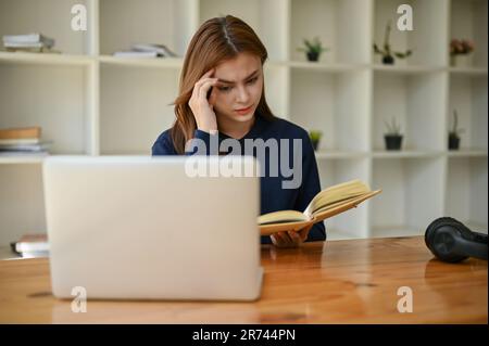 Eine gestresste und ernsthafte junge asiatische Studentin konzentriert sich darauf, ein Buch an ihrem Schreibtisch in der Bibliothek zu lesen. Bildungskonzept Stockfoto