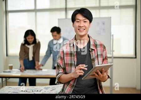 Ein attraktiver und cleverer junger asiatischer Websiteentwickler in Flanellhemden steht im Büro mit einem digitalen Tablet in der Hand, während seine Kollegen A haben Stockfoto