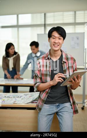 Das Porträt eines gutaussehenden und klugen jungen asiatischen Websiteentwicklers für Freizeitkleidung steht im Büro mit einem digitalen Tablet in der Hand, während h Stockfoto