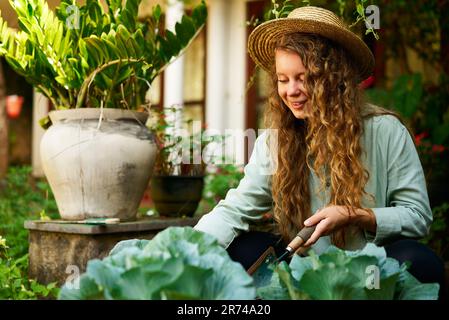 Junge, weiße Gärtnerin, die Gemüse pflanzt und ein glückliches Lächeln aus der Nahaufnahme macht. Junge, fröhliche Frau mit Kelle in ihrem Garten, die sich um sie kümmert Stockfoto
