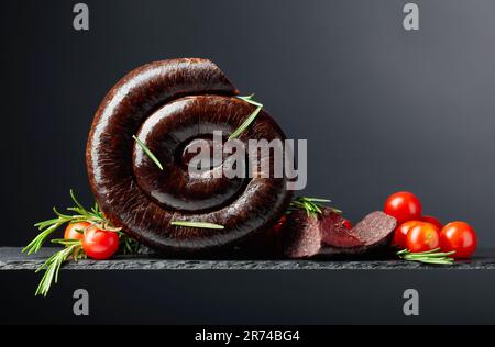 Traditioneller europäischer schwarzer Pudding oder Blutwurst mit Rosmarin und Tomaten auf schwarzem Hintergrund. Speicherplatz kopieren. Stockfoto