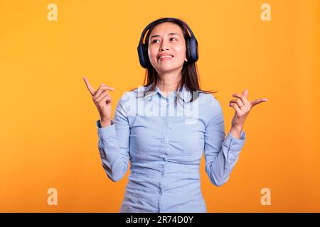 Lächelnde positive Frau, die im Studio Musik hört und Tanzbewegungen auf gelbem Hintergrund zeigt. Fröhliches philippinisches Model mit Kopfhörern, das zu ihrem Lieblingslied tanzt Stockfoto