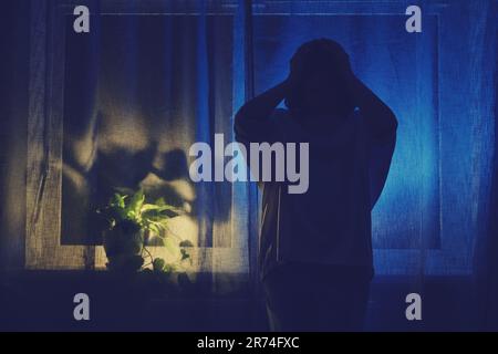 Der Schatten einer weiblichen Figur hält ihren Kopf mit den Händen gegen den Hintergrund eines Fenstervorhangs. Eine Frau trauert um den dunklen Wind der Nacht Stockfoto