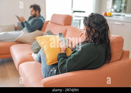 Ein junges Ehepaar, das auf einer Couch in einem sonnenbeleuchteten Wohnzimmer sitzt. Sie sind in ihre Smartphones vertieft, ohne sich der Gegenwart des anderen bewusst zu sein. Negativ i Stockfoto