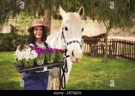 Frau in Arbeitskleidung, Handschuhen und Strohhut, die mit Blumen im Garten arbeitet, weißes Pferd in der Nähe, verschwommene grüne Bäume und Gartenhintergrund Stockfoto