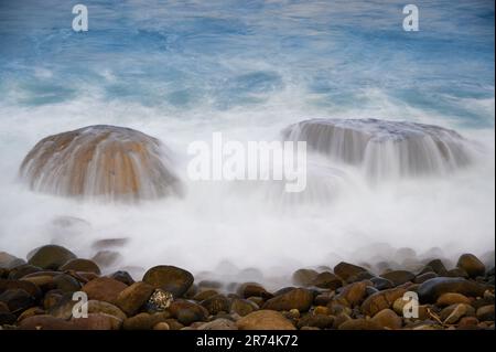 Weiße Wellen fließen langsam von den Felsen ins Meer. Aufnahme mit geringer Belichtung. Blick auf Keelung Harbor vom Wanli Beach in New Taipei City. Taiwan. Stockfoto
