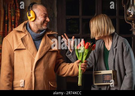 Lustiges Foto eines Mannes mit Kopfhörern aus dem Lärm, der einer Frau mit einem Retro-Radio einen Strauß Tulpen gibt. Stockfoto
