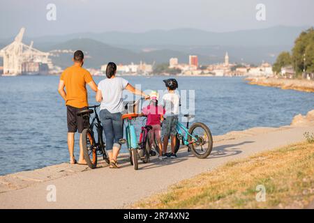 Zwei Kinder mit Helmen auf dem Kopf und lächelnde Eltern, die auf einer familienfreundlichen Radroute entlang der Küste mit Blick auf die Küste Fahrräder fahren. Stockfoto