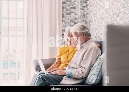 Ziemlich ältere 70s-jährige, grauhaarige Paar, das sich im Wohnzimmer auf dem Sofa ruht, halten Sie den Laptop, und schauen Sie sich einen lächelnden Film an. Genießen Sie Freizeit, ältere Generationen und moderne Wireless-Technologie fortschrittliches Benutzerkonzept Stockfoto
