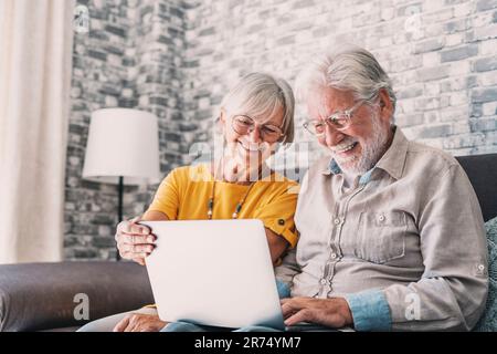 Ziemlich ältere 70s-jährige, grauhaarige Paar, das sich im Wohnzimmer auf dem Sofa ruht, halten Sie den Laptop, und schauen Sie sich einen lächelnden Film an. Genießen Sie Freizeit, ältere Generationen und moderne Wireless-Technologie fortschrittliches Benutzerkonzept Stockfoto