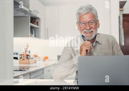 Ein alter Telearbeiter. Ein konzentrierter Seniorenmännchen mit Brille arbeitet auf einem Laptop vom Heimbüro aus und schaut auf die Kamera. Freiberuflicher Mitarbeiter im Alter sitzt am Küchentisch und schreibt den Bericht online Stockfoto