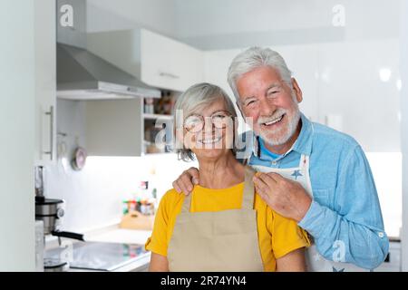 Kopfschuss Porträt lächelndes, reifes Ehepaar, das in die Kamera schaut, in der Küche steht und zu Hause kocht, glücklicher Seniorenmann mit Brille umarmt Ehefrau, Großeltern posieren für ein Foto Stockfoto
