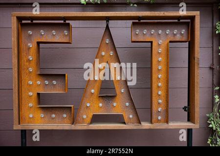 Großes, rechteckiges Schild mit DER Aufschrift EAT, das Glühbirnen hat und rostig ist, steht vor einer braun bemalten Holzplankenwand. Stockfoto