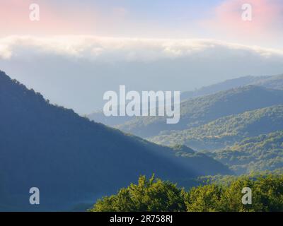 Blick auf grüne bergige bewaldete Hügel im Nebel. Dramatische Wolken über dem fernen Kamm Stockfoto