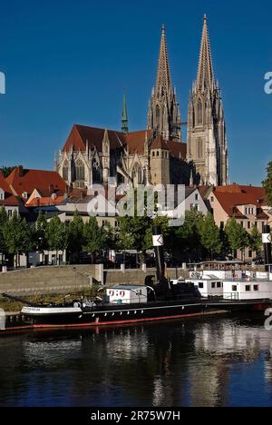 Die zwei neogotischen Türme des Petersdoms, die in den 1800er Jahren in einem überwiegend mittelalterlichen Gebäude aus den Jahren 1275 bis 1520 errichtet wurden, ragten über der Donau in Regensburg in Deutschland. Unter der Kathedrale, am Fluss, befindet sich ein historisches Dampfboot aus dem Jahr 1923, die Ruthof. Stockfoto