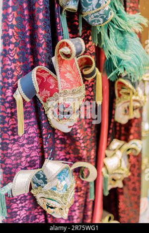 Venezianische Masken hängen an der Decke. Im Hintergrund sind Stoffe in verschiedenen Farben zu sehen. Die Masken sind farbenfroh und bereit für einen Maskenball. Stockfoto