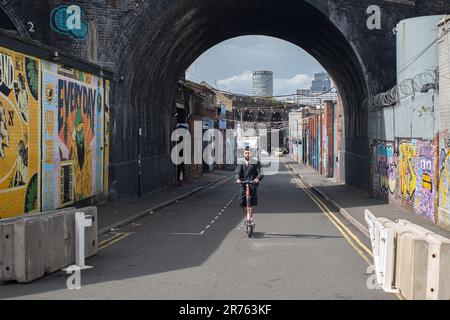 Ein Mann fährt mit einem elektrischen Roller mitten auf der Straße in einer rauen städtischen Umgebung. Eisenbahnbögen und ein Stadtbild sind hinter ihm zu sehen. Stockfoto