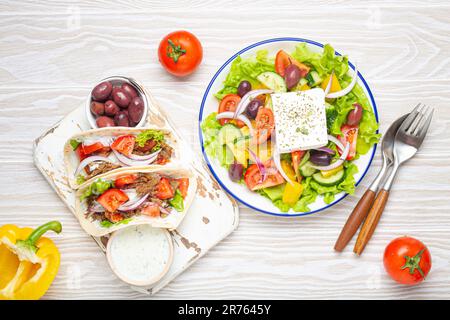 Traditionelle griechische Speisen: Griechischer Salat, Gyros mit Fleisch und Gemüse, Tzatziki-Sauce, Oliven auf weißem rustikalem Holztisch von oben Stockfoto