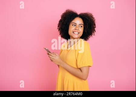 Hübsche afroamerikanische oder brasilianische Frau, in gelbem Kleid, mit ihrem Smartphone, im Internet mit Freunden chatten, im Internet surfen, SMS schreiben, wegschauen, lächeln, Stellen Sie sich auf einen isolierten rosa Hintergrund Stockfoto