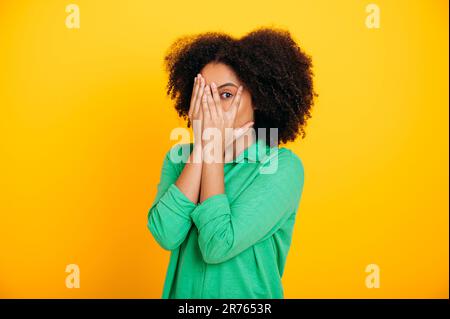 Beängstigend, ängstlich. Verängstigte hispanische oder brasilianische lockige Frau in einem legeren Hemd, schaut durch die Finger, bedeckt das Gesicht mit Händen, erfährt Angst, Schock, steht auf isoliertem gelben Hintergrund Stockfoto